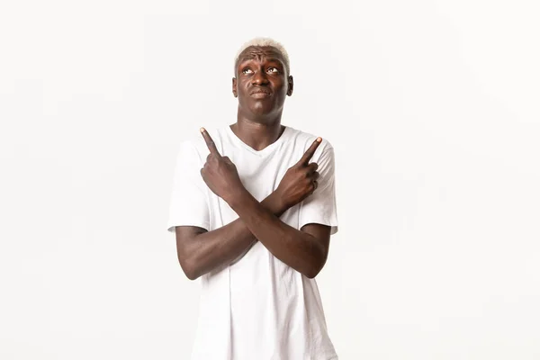 Портрет нерешительного африканско-американского парня, принимающего решение, указывающего пальцем в сторону и выглядящего смущенным, стоящего на белом фоне — стоковое фото