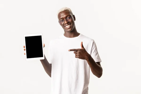 Retrato de atractivo chico afroamericano sonriente en gafas que apuntan con el dedo a la pantalla de la tableta digital, recomiendan aplicación, mostrando algo, de pie fondo blanco — Foto de Stock