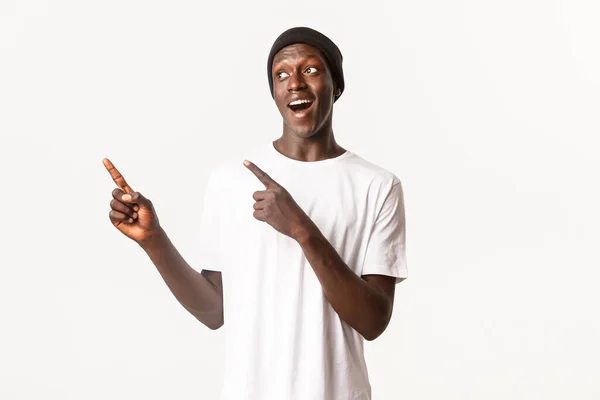 Portré izgatott és elégedett afro-amerikai srác sapkában, ujjal mutogató és keres balra érdekes kifejezés, fehér háttér — Stock Fotó