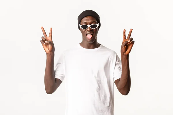 Портрет смешного и жизнерадостного африканско-американского молодого человека в шапочках и солнцезащитных очках, показывающего жесты мира и прилипающий язык, белый фон — стоковое фото