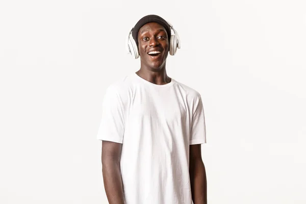 Retrato de un joven afroamericano divertido y feliz escuchando podcast o música en auriculares, sonriendo contento, fondo blanco — Foto de Stock