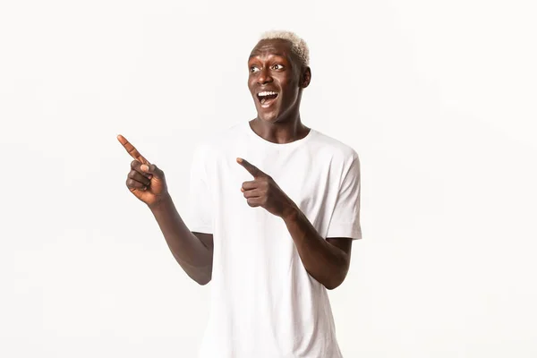 Retrato de animado cara loiro afro-americano bonito, olhando feliz e surpreso com o logotipo, apontando os dedos canto superior esquerdo, fundo branco — Fotografia de Stock
