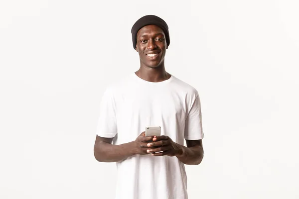 Portré vidám afro-amerikai modern srác sapkában, kezében mobiltelefon és mosolygós boldog a kamera, alkalmazás, fehér háttér — Stock Fotó