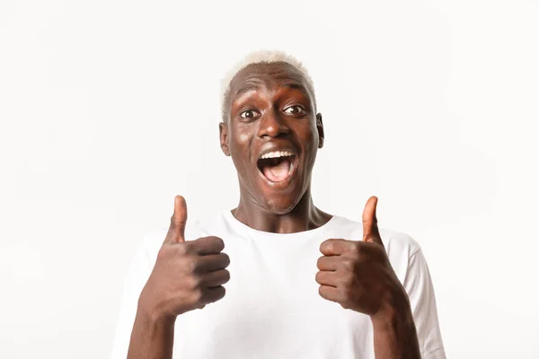 Retrato de alegre e animado cara loiro afro-americano bonito olhando com alegria, mostrando polegares para cima e sorrindo, aprovar ou recomendar algo — Fotografia de Stock