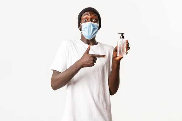 Πορτρέτο του ενθουσιασμένος όμορφος και κομψός αφροαμερικανός τύπος σε beanie και ιατρική μάσκα, δείχνοντας δάχτυλο στο χέρι μπουκάλι απολυμαντικό, στέκεται λευκό φόντο — Φωτογραφία Αρχείου