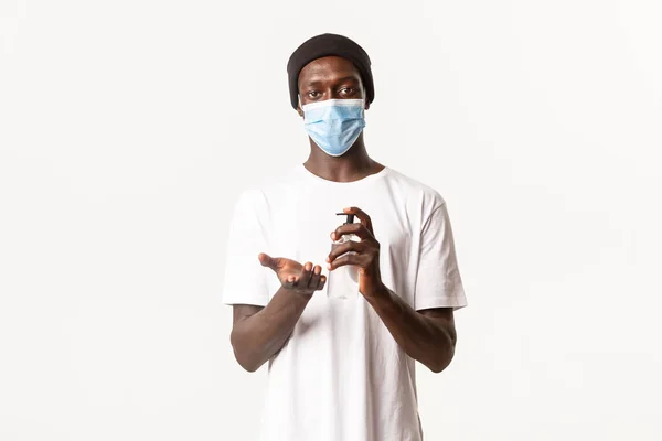 Jonge Afrikaans-Amerikaanse man met medisch masker, met behulp van het voorkomen van maatregelen van covid-19, het toepassen van de hand sanitizer op handen, staande over witte achtergrond — Stockfoto