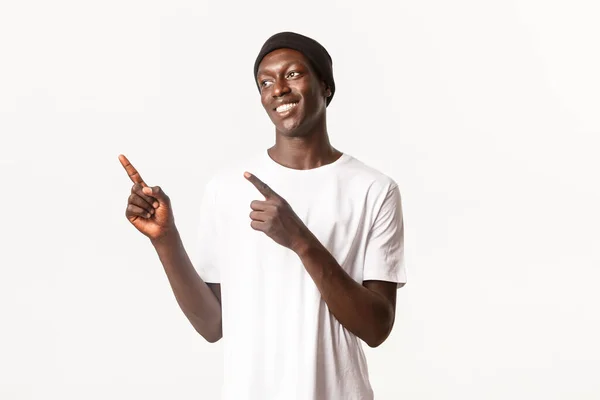 Portrét šťastného pohledného afroamerického mladíka, oblečený v korále a tričku, s potěšením se usmívá a dívá se, ukazuje na levý horní roh, bílé pozadí — Stock fotografie