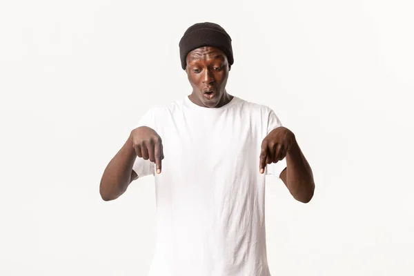 Retrato de chico afroamericano maravillado y sorprendido en gorro, hombre guapo señalando con los dedos hacia abajo y mirando sorprendido, de pie fondo blanco — Foto de Stock