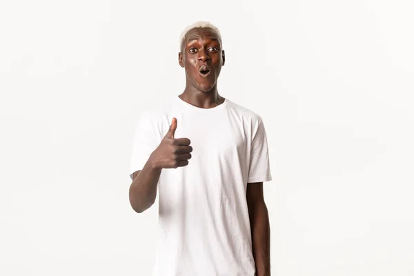 Retrato de homem loiro afro-americano impressionado, sorrindo espantado e mostrando o polegar para cima na aprovação, como algo, fundo branco — Fotografia de Stock