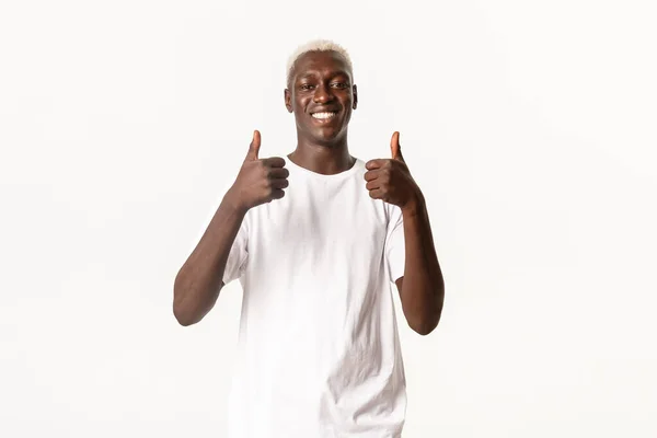 Retrato de sorrir cara afro-americano bonito, parecendo satisfeito, mostrando polegar-se em aprovação, como algo, fundo branco — Fotografia de Stock