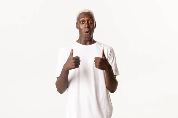 Retrato de homem loiro afro-americano maravilhado e impressionado, mostrando polegares para cima fascinado, de pé fundo branco — Fotografia de Stock