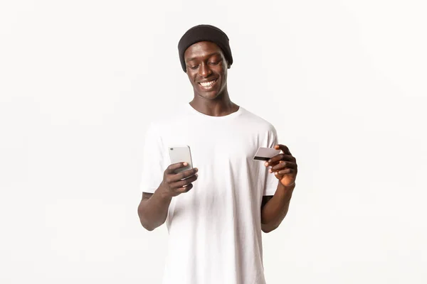 Retrato del guapo hombre afroamericano sonriente comprando en línea, con tarjeta de crédito y teléfono móvil, fondo blanco — Foto de Stock