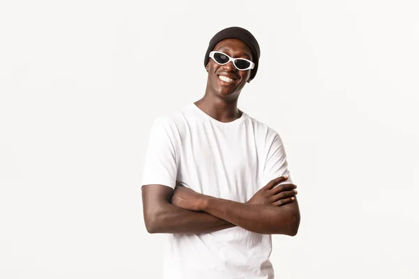 Retrato de chico afroamericano atrevido y elegante en gorro, gafas de sol, de pie satisfecho con los brazos cruzados y sonriendo feliz ante la cámara, fondo blanco — Foto de Stock