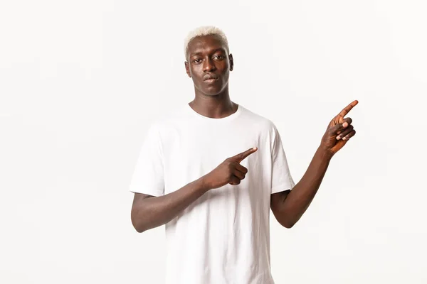 Портрет серьезного африканско-американского блондина, указывающего пальцем в правом верхнем углу и показывающего логотип, стоящего на белом фоне — стоковое фото