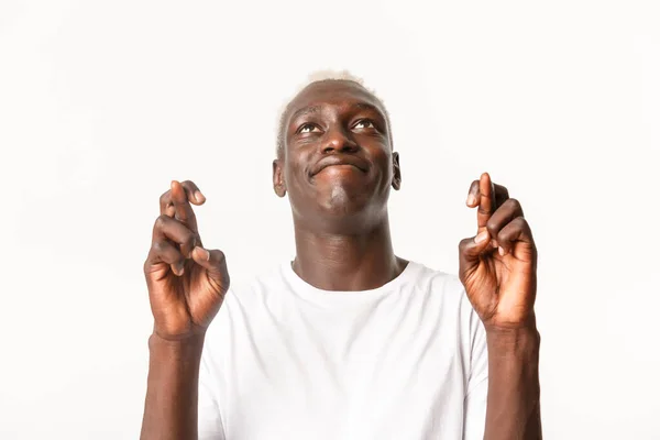 Primo piano di speranzoso giovane uomo afro-americano con i capelli biondi, che alza lo sguardo verso il cielo e prega, implora Dio mentre esprime desideri con le dita incrociate, in piedi sfondo bianco — Foto Stock