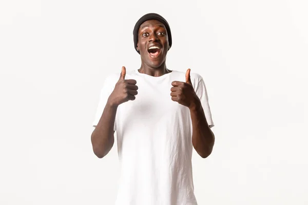 ビーニーで興奮し陽気なアフリカ系アメリカ人の男の肖像画、面白いと承認で親指アップを示す、白い背景 — ストック写真
