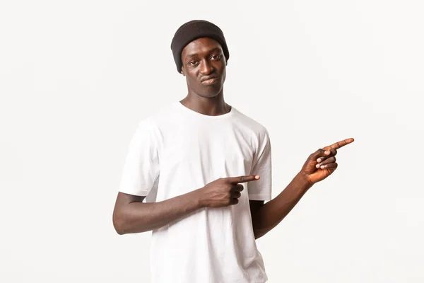 Retrato de chico afroamericano escéptico y crítico en gorro, señalando con los dedos hacia la derecha y con muecas decepcionados, expresan aversión, de pie fondo blanco — Foto de Stock