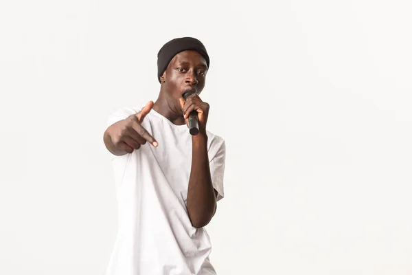 Portrait of sassy and cool Black rapper in beanie, singing into microphone and making hip-hop gesture, white background — Stock Photo, Image