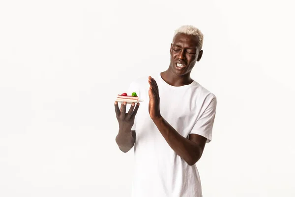 うんざりハンサムなアフリカ系アメリカ人の若いブロンドの男の肖像画、不本意ながらデザートを食べる、嫌なケーキからくさび、白い背景に立って — ストック写真