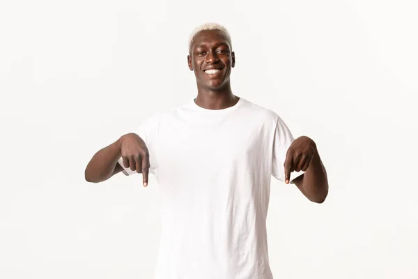 Portrait of stylish handsome african american blond man, smiling satisfied and pointing fingers down, white background — Stock Photo, Image