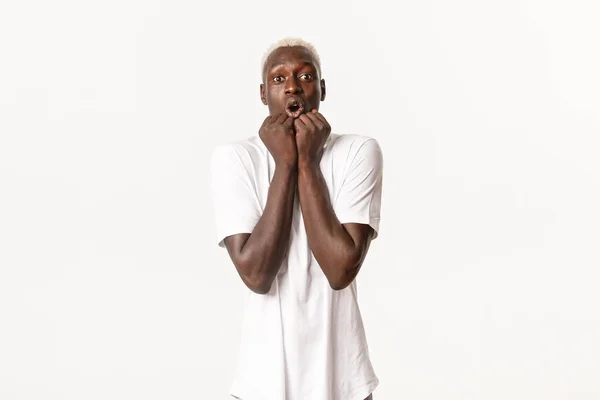 Portrait of amazed and fascinated african-american blond man, gasping excited, holding hands pressed to face, looking at camera thrilled, standing white background — Stock Photo, Image