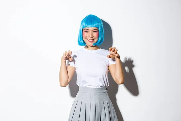 Chica asiática divertida y linda en peluca azul sonriendo y haciendo el gesto de las patas de la mano, celebrando halloween, de pie sobre fondo blanco — Foto de Stock