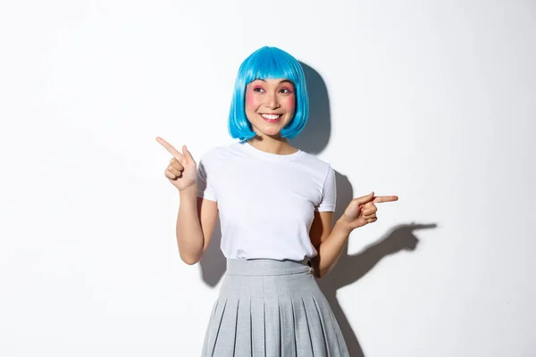Retrato de mujer sonriente asombrada en peluca azul vestida para la celebración de Halloween, señalando los dedos hacia los lados, mostrando dos promociones, de pie sobre fondo blanco — Foto de Stock