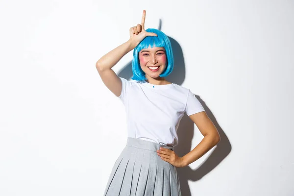 Retrato de bela menina asiática boba celebrando halloween, mostrando gesto perdedor na testa e sorrindo, zombando de alguém, de pé sobre fundo branco — Fotografia de Stock