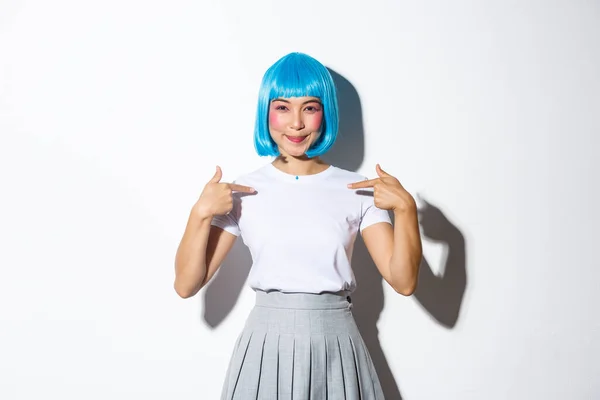 Hermosa chica asiática en peluca azul señalándose los dedos a sí misma y sonriendo, de pie confiada sobre el fondo blanco, vistiendo traje de halloween — Foto de Stock
