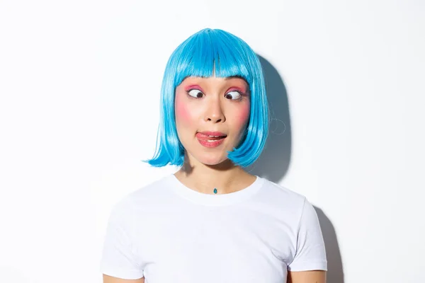 Крупный план смешной и глупой азиатской девушки-артистки, празднующей Хэллоуин, носящей синий парик и щурящейся, показывающей гримасы — стоковое фото