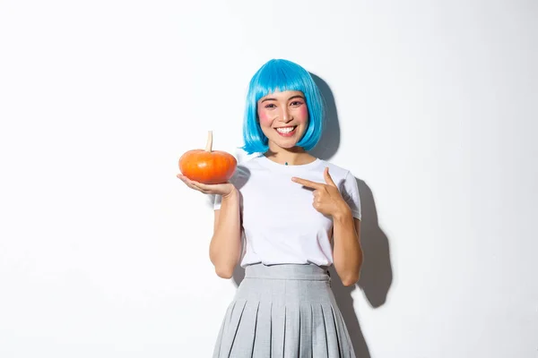 Carino sorridente donna asiatica in parrucca blu che punta il dito alla piccola zucca, celebrando Halloween, in piedi su sfondo bianco — Foto Stock