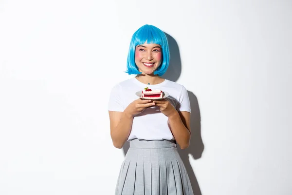 Retrato de hermosa chica asiática sonriente en peluca de anime azul sonriendo y mirando a la izquierda, sosteniendo el plato con pastel, de pie sobre fondo blanco — Foto de Stock