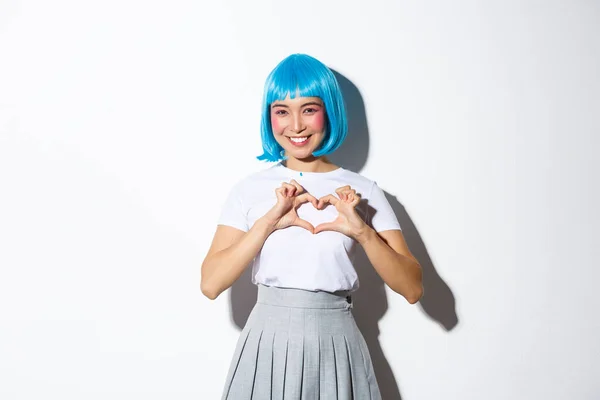 Retrato de hermosa chica asiática mirando kawaii y mostrando gesto de corazón, sonriendo mientras está de pie sobre fondo blanco en traje de halloween y peluca azul — Foto de Stock