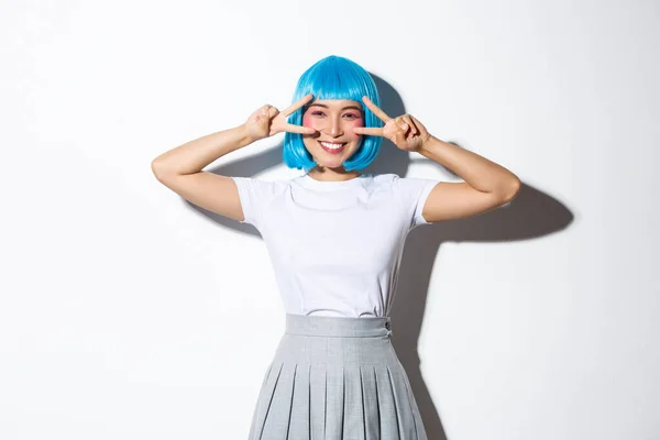 Alegre bonita asiática mujer en azul fiesta peluca, celebrando evento, mostrando gestos de paz y sonriendo feliz, de pie sobre fondo blanco — Foto de Stock