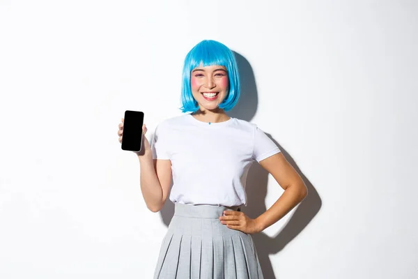 Portrait of cute asian girl dressed as anime character in blue wig, smiling happy and showing smartphone screen, standing over white background