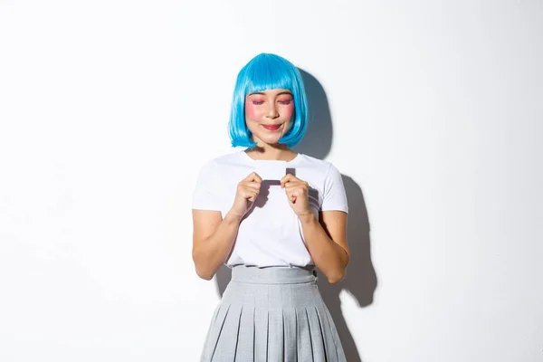 Portret pięknej azjatyckiej dziewczyny w niebieskiej peruce i kostiumie halloween, marzącej o zakupach trzymając kartę kredytową i uśmiechając się zamkniętymi oczami, stojącej na białym tle — Zdjęcie stockowe