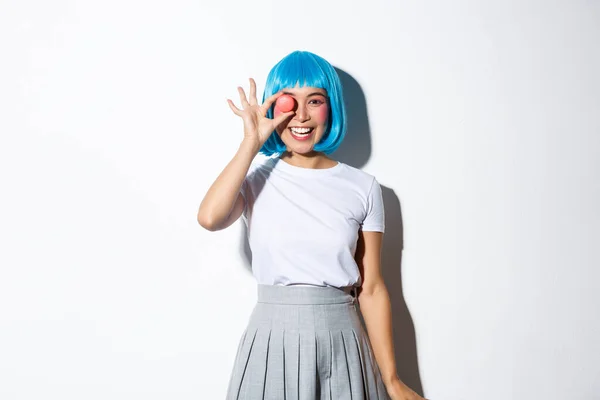 Linda chica asiática en peluca de anime y uniforme de colegiala mirando feliz, mostrando macaron y sonriendo, de pie sobre fondo blanco — Foto de Stock