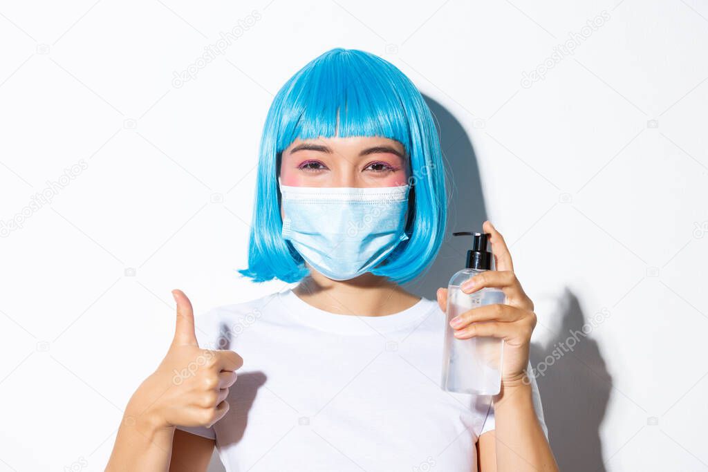 Concept of halloween celebration and coronavirus. Close-up of cute smiling asian girl in blue wig and medical mask showing hand saniziter and thumb-up