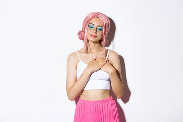 Πορτρέτο της νοσταλγικής όμορφη νεαρή γυναίκα σε ροζ περούκα, φωτεινό μακιγιάζ, κλείστε τα μάτια και κρατώντας τα χέρια στην καρδιά, να θυμάστε κάτι ή ονειροπόληση, στέκεται πάνω από το λευκό φόντο — Φωτογραφία Αρχείου