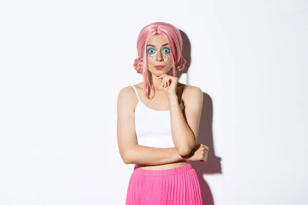 Podekscytowany cute party girl z różową peruką i makijaż halloween, patrząc z zainteresowaniem przed kamerą, słuchając czegoś, stojąc na białym tle — Zdjęcie stockowe
