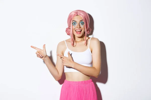 Retrato de menina feliz animado em peruca rosa e maquiagem brilhante celebrando algo, apontando os dedos deixados para o seu logotipo sobre feriados e festas, de pé sobre fundo branco — Fotografia de Stock