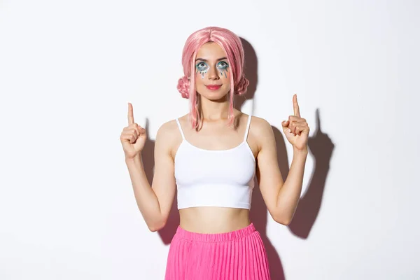 Retrato de menina bonita festa com peruca rosa e maquiagem glamour, apontando os dedos e olhando para cima, mostrando seu banner logotipo, de pé sobre fundo branco — Fotografia de Stock