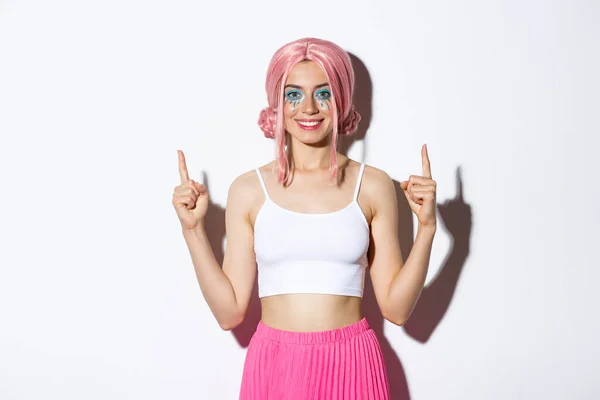 Portret uśmiechniętej modelki z peruką imprezową i makijażem halloween, wskazującym palcem w górę, pokazującym logo, stojącym nad białym tłem — Zdjęcie stockowe