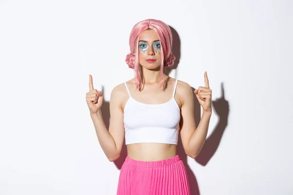 Retrato de menina de festa elegante com peruca de anime rosa e maquiagem halloween, apontando os dedos para o seu logotipo, de pé sobre fundo branco — Fotografia de Stock