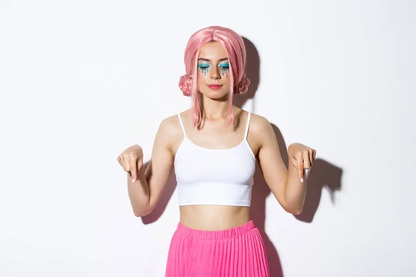 Retrato de menina festa sorridente em peruca rosa e roupa para o dia das bruxas, apontando e olhando para baixo para o seu logotipo, de pé sobre fundo branco — Fotografia de Stock