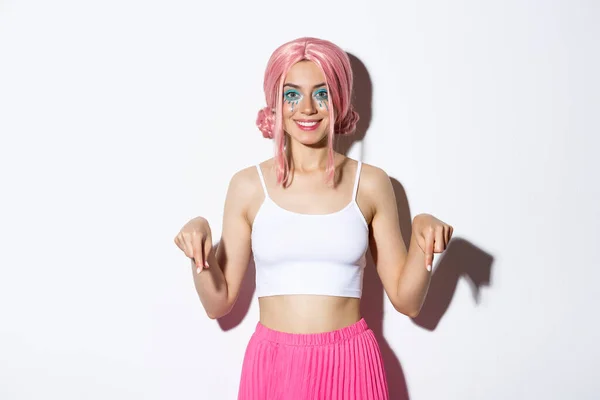 Piękny uśmiech dziewczyna strona z różowymi włosami i jasny makijaż, wskazując palcami w dół na logo, noszenie kostiumu halloween, stojąc na białym tle — Zdjęcie stockowe