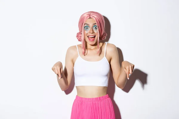 Porträt eines aufgeregten Partygirls in rosa Perücke und Halloween-Outfit, das amüsiert und glücklich aussieht, während es mit dem Finger nach unten zeigt, vor weißem Hintergrund stehend — Stockfoto