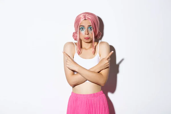 Αναποφάσιστη ανόητο θηλυκό μοντέλο σε ροζ περούκα, με πολύχρωμο μακιγιάζ για αποκριές κόμμα, δείχνοντας πλάγια και shrugging, ζητώντας βοήθεια με την επιλογή, στέκεται ανίδεος πάνω από λευκό φόντο — Φωτογραφία Αρχείου