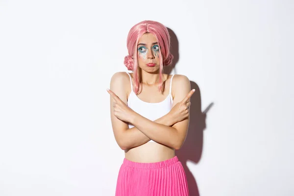 Portrait of clueless silly young girl in pink wig and halloween costume, shrugging and pouting indecisive, pointing fingers sideways, standing over white background — Stock Photo, Image