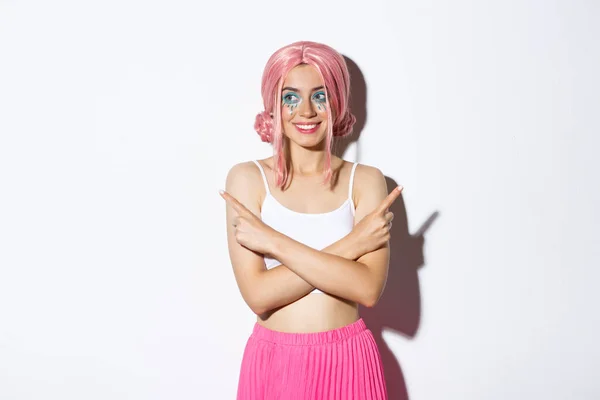 Menina bonita festa sorridente em traje de Halloween e peruca rosa, apontando os dedos para o lado, mas olhando para a esquerda, fazendo escolha, de pé sobre fundo branco — Fotografia de Stock